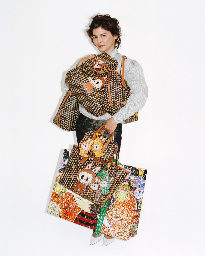 Audrey Tautou standing against a white backdrop holding an array of Moynat monogram tote bags featuring character art.