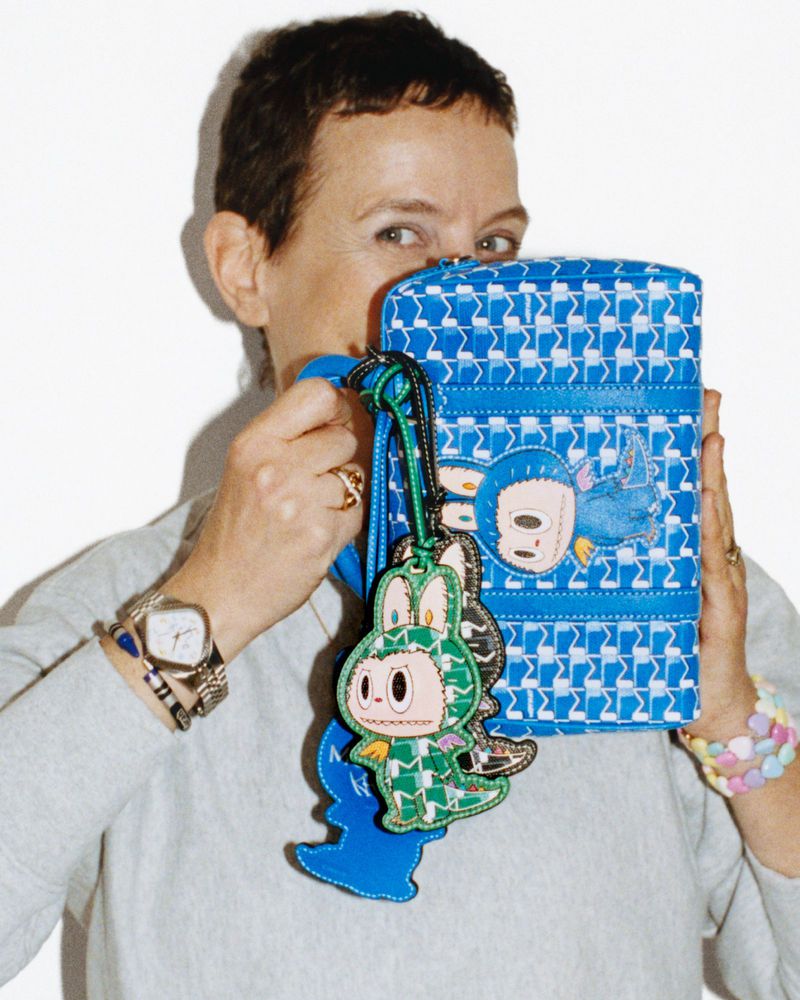 Woman with short hair holding a blue patterned Moynat handbag with monster charms in front of her face against a white backdrop.