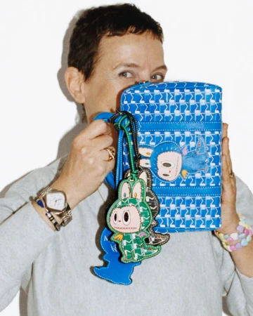 Woman with short hair holding a blue patterned Moynat handbag with monster charms in front of her face against a white backdrop.