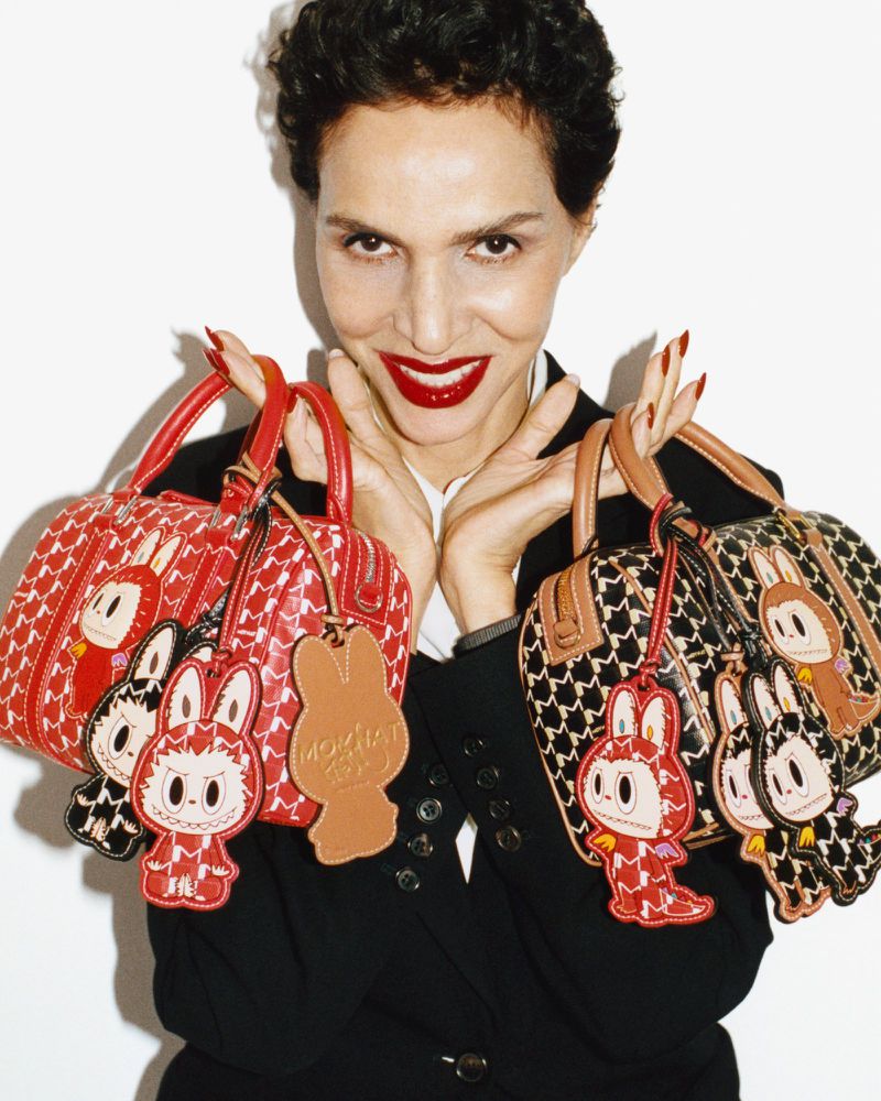 Farida Khelfa smiling while holding two patterned Moynat handbags adorned with character charms against a white background.