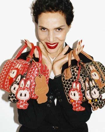 Farida Khelfa smiling while holding two patterned Moynat handbags adorned with character charms against a white background.