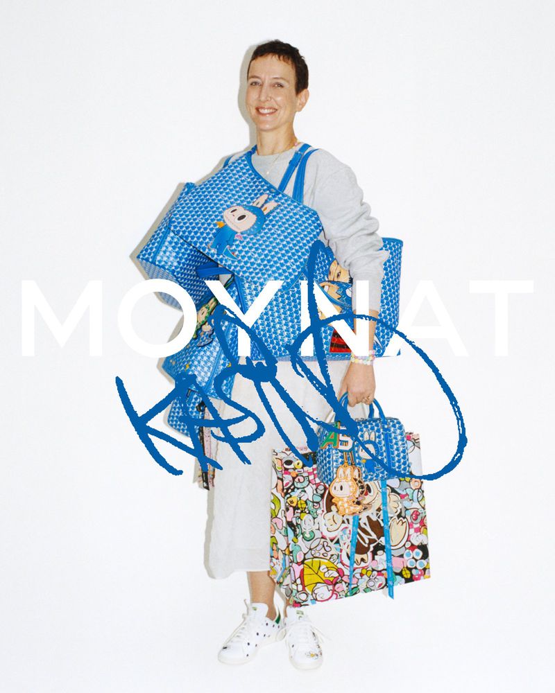 Sarah Andelman holding printed blue Moynat x Kasing Lung monogram tote bags against a white seamless studio backdrop.