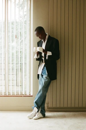 Model Anyier Anei leaning against an interior wall wearing an oversized black blazer, white gloves, and blue jeans beside a tall window.