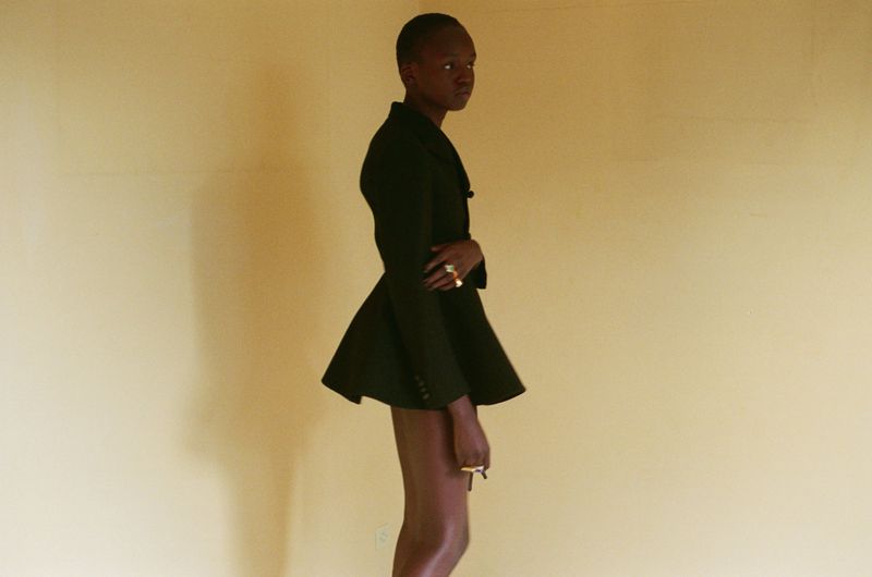 Model Anyier Anei standing in profile wearing a structured black flared jacket against a warm beige background.