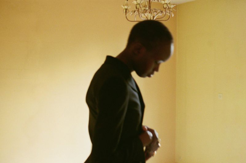 Model Anyier Anei captured in profile wearing a dark tailored blazer against a warm yellow interior wall beneath a chandelier.