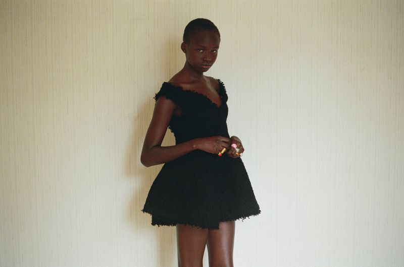 Model Anyier Anei wearing an off-the-shoulder black tweed mini dress against a textured neutral wall.