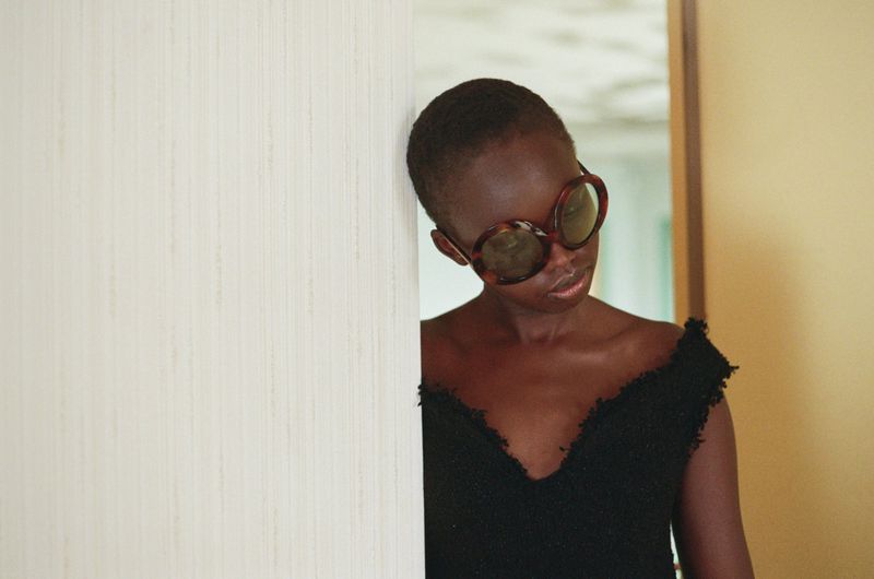 Model Anyier Anei wearing oversized tortoiseshell sunglasses and a black textured top leaning against a wall