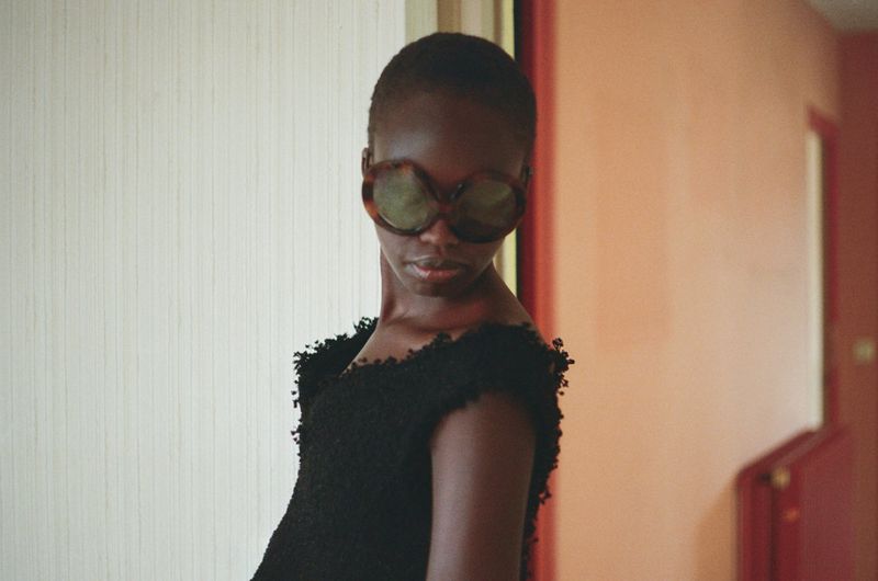 Model Anyier Anei wearing large round sunglasses and a textured black sleeveless garment in a hallway.