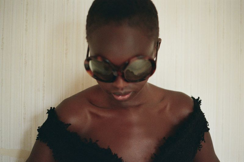 Anyier Anei wearing oversized tortoiseshell sunglasses and a black off-the-shoulder textured top against a neutral wall.