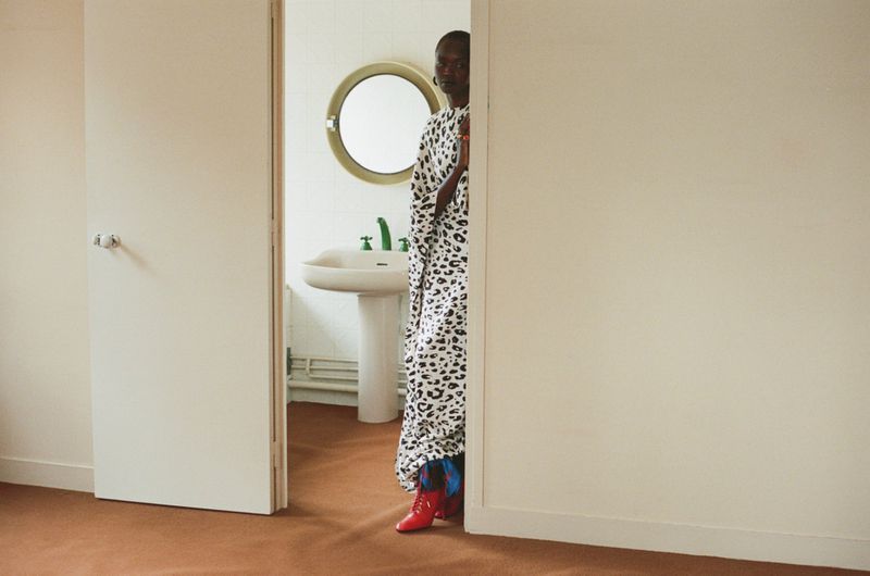 Model Anyier Anei wearing a leopard print dress and red boots, standing partially behind a door leading to a bathroom.