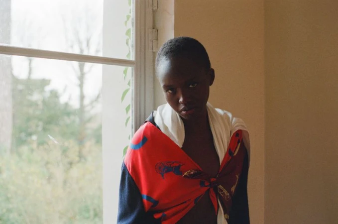 Model Anyier Anei wearing a tied red printed silk scarf and looking toward the camera beside an interior window.