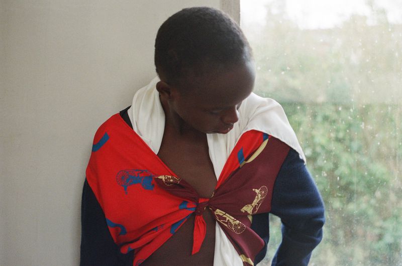 Model Anyier Anei looking down, styled in a tied red printed scarf and navy sleeve by a window.