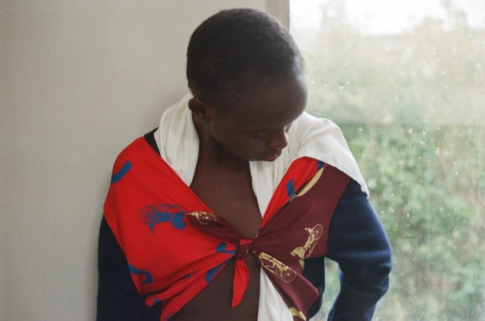 Model Anyier Anei looking down, styled in a tied red printed scarf and navy sleeve by a window.