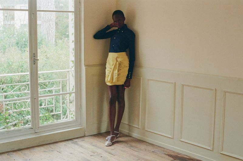 Anyier Anei wearing a dark navy button-up shirt and a draped yellow skirt, leaning against a wall in an interior room.