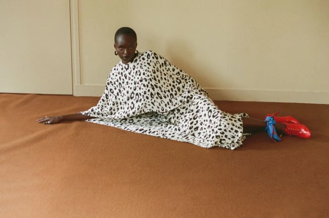 Model Anyier Anei lying on a brown carpeted floor wearing a black and white leopard print draped dress and red heels.