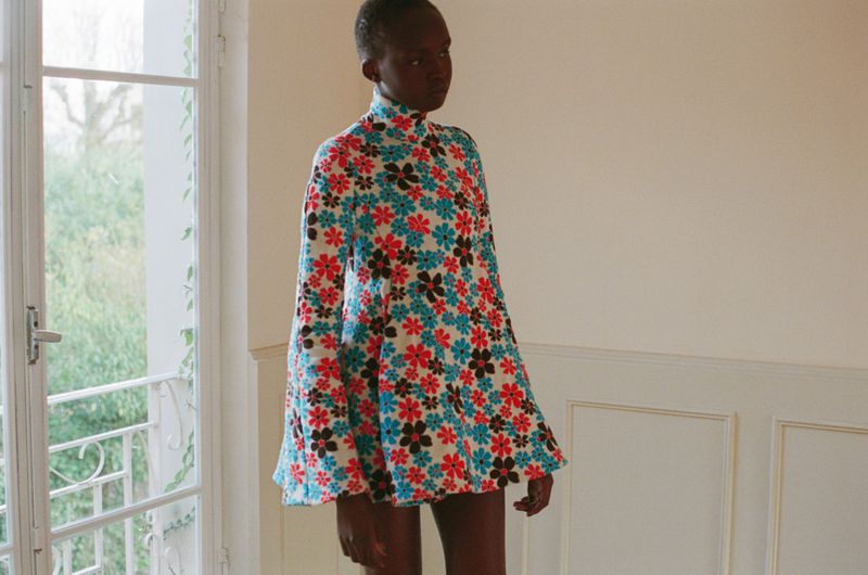 Model Anyier Anei poses in a high-neck floral mini dress with bell sleeves beside an open French window.