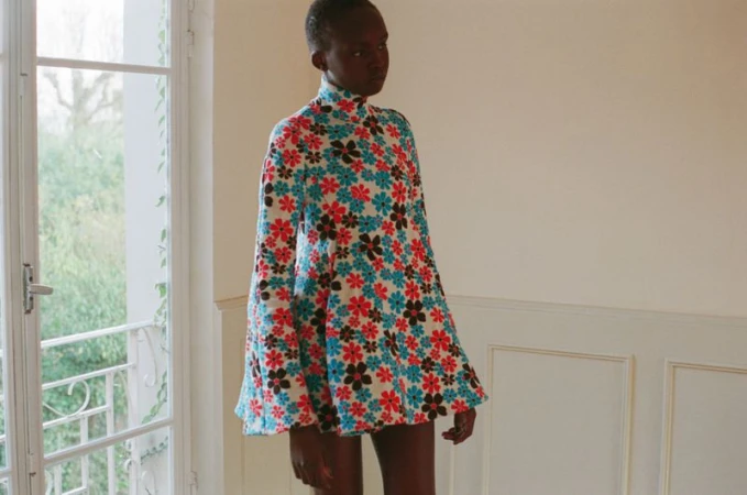 Model Anyier Anei poses in a high-neck floral mini dress with bell sleeves beside an open French window.