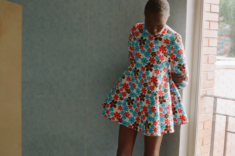 Model Anyier Anei wearing a floral print mini dress with long sleeves while posing indoors next to a window.