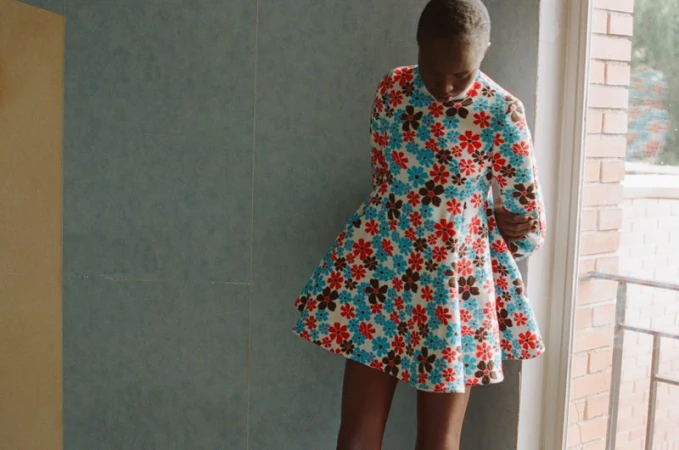 Model Anyier Anei wearing a floral print mini dress with long sleeves while posing indoors next to a window.