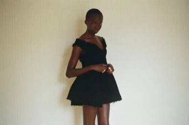 Model Anyier Anei wearing an off-the-shoulder black tweed mini dress against a textured neutral wall.