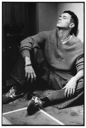 Black and white photo of actor Arón Piper seated on the floor leaning against a wall wearing a Dior sweater and sneakers.