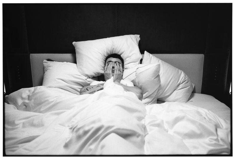 Actor Arón Piper lying in a bed surrounded by white pillows with hands over his face in a black and white portrait.