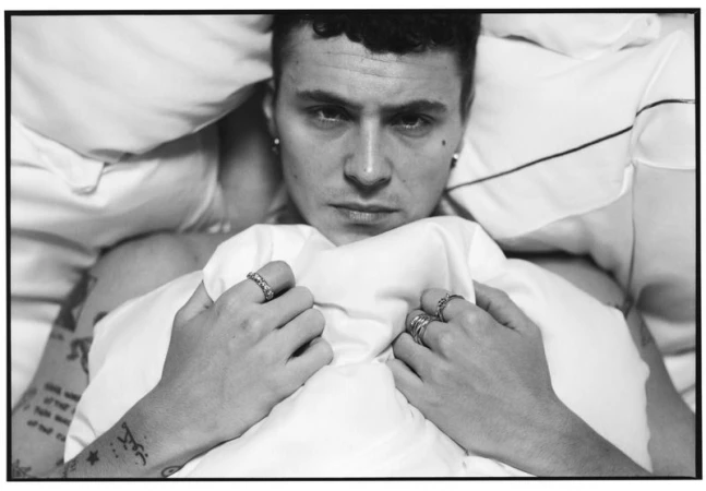 Close-up monochrome portrait of actor Ar3n Piper lying in bed with white pillows, holding a duvet up with rings on his fingers.