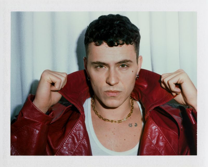 Actor Arón Piper wearing a red leather jacket and gold chain necklace against a pleated fabric backdrop.
