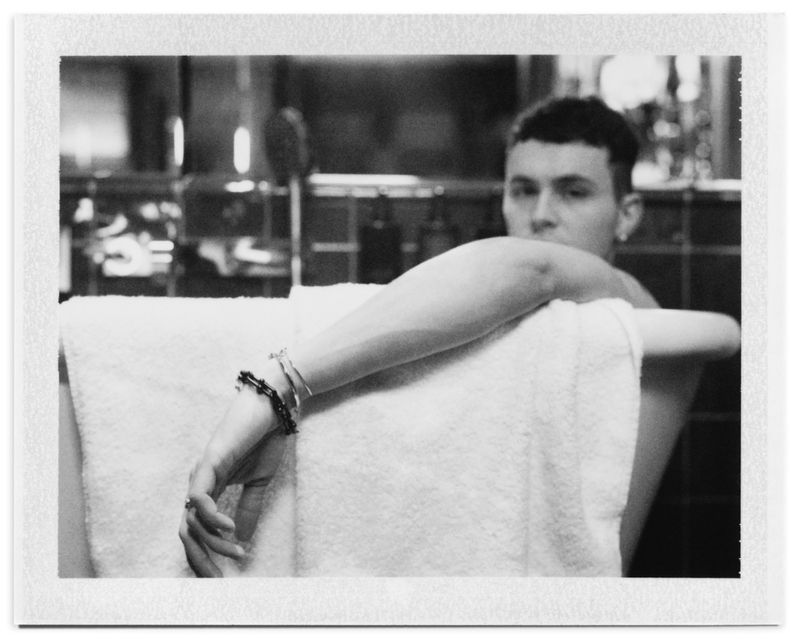 Black and white portrait of Arón Piper resting his arm over a white towel draped on a bathtub in a bathroom.