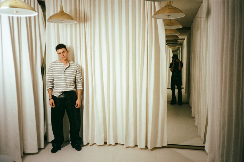 Arón Piper wearing a striped long-sleeve shirt and black wide-leg trousers posing in front of cream pleated curtains.