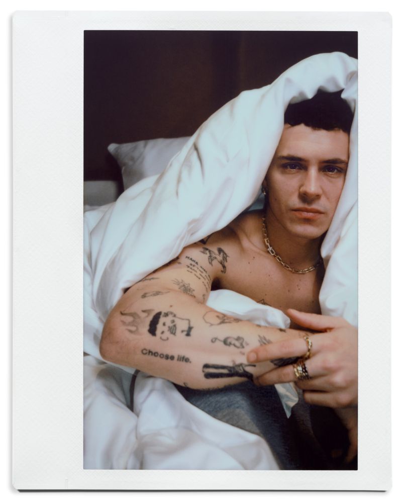 Shirtless Arón Piper draped in a white duvet in bed showing arm tattoos and gold jewelry in a polaroid portrait.