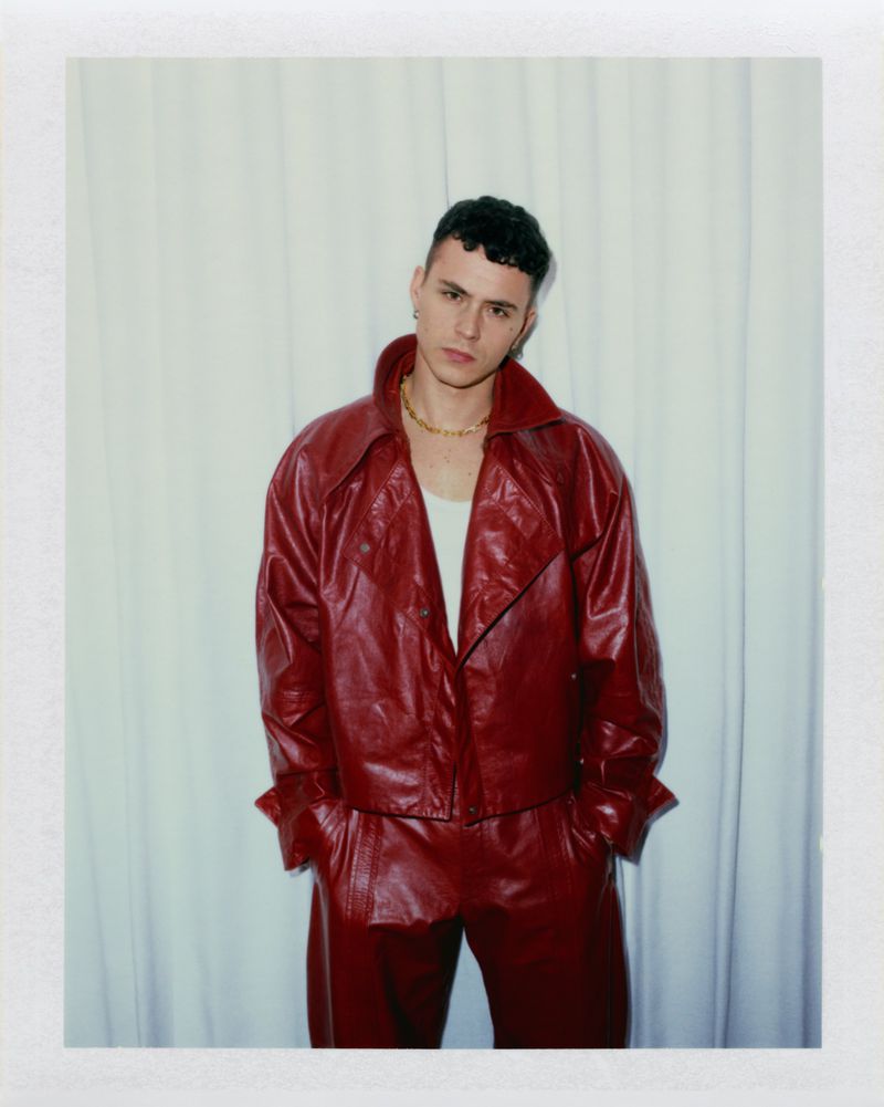 Actor Arón Piper wearing a red leather jacket and matching trousers against a draped white curtain in a Polaroid portrait.