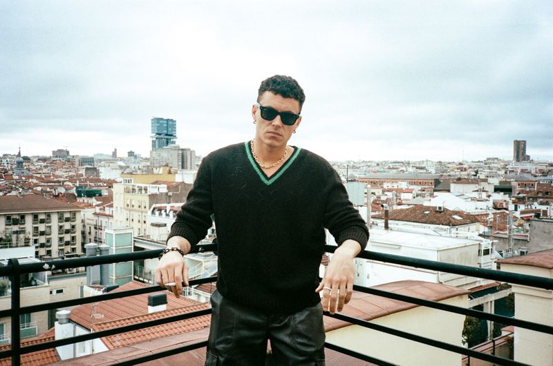 Arón Piper wearing sunglasses and a black knit v-neck sweater leaning on a rooftop railing overlooking a city.