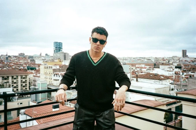 Arón Piper wearing sunglasses and a black knit v-neck sweater leaning on a rooftop railing overlooking a city.