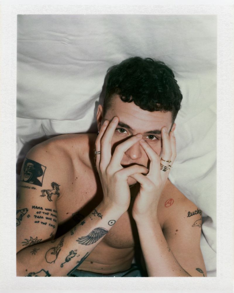 Polaroid portrait of actor Arón Piper shirtless with arm tattoos and rings covering his face against white bedding.