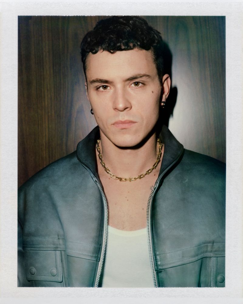 Actor Arón Piper wearing a slate blue leather jacket and gold chain necklace in a Polaroid portrait against a wood panel wall.