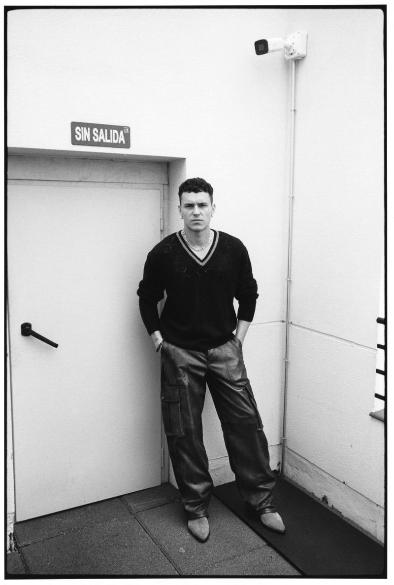 Actor Aron Piper posing outside a door with a SIN SALIDA sign in a black V-neck sweater and dark trousers for Behind the Blinds.