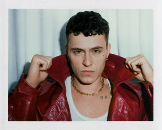 Actor Arón Piper wearing a red leather jacket and gold chain necklace against a pleated fabric backdrop.