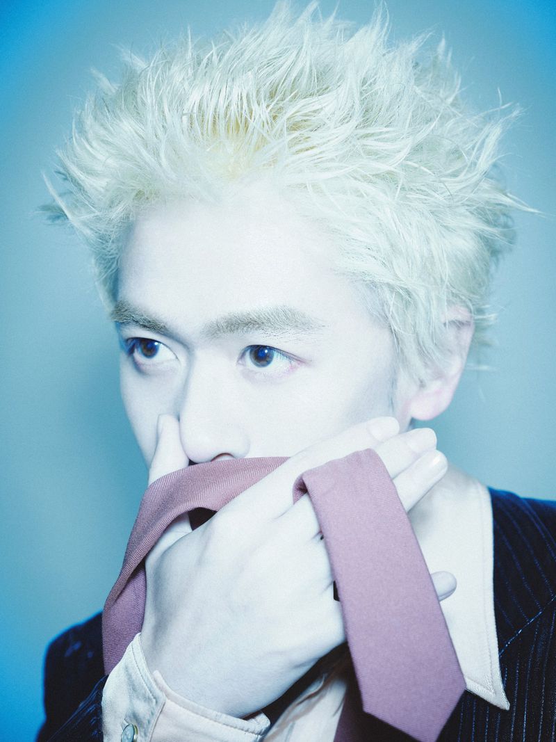 Close-up portrait of an East Asian man with spiky bleached blond hair holding a mauve tie across his face against a blue background.