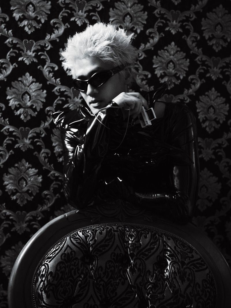 Man with spiky bleached hair wearing wraparound black sunglasses and a glossy vinyl top against ornate damask wallpaper.
