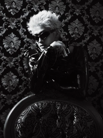 Man with spiky bleached hair wearing wraparound black sunglasses and a glossy vinyl top against ornate damask wallpaper.