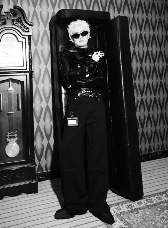 Model in sunglasses and wide-leg trousers standing inside an open leather case beside a grandfather clock in black-and-white.