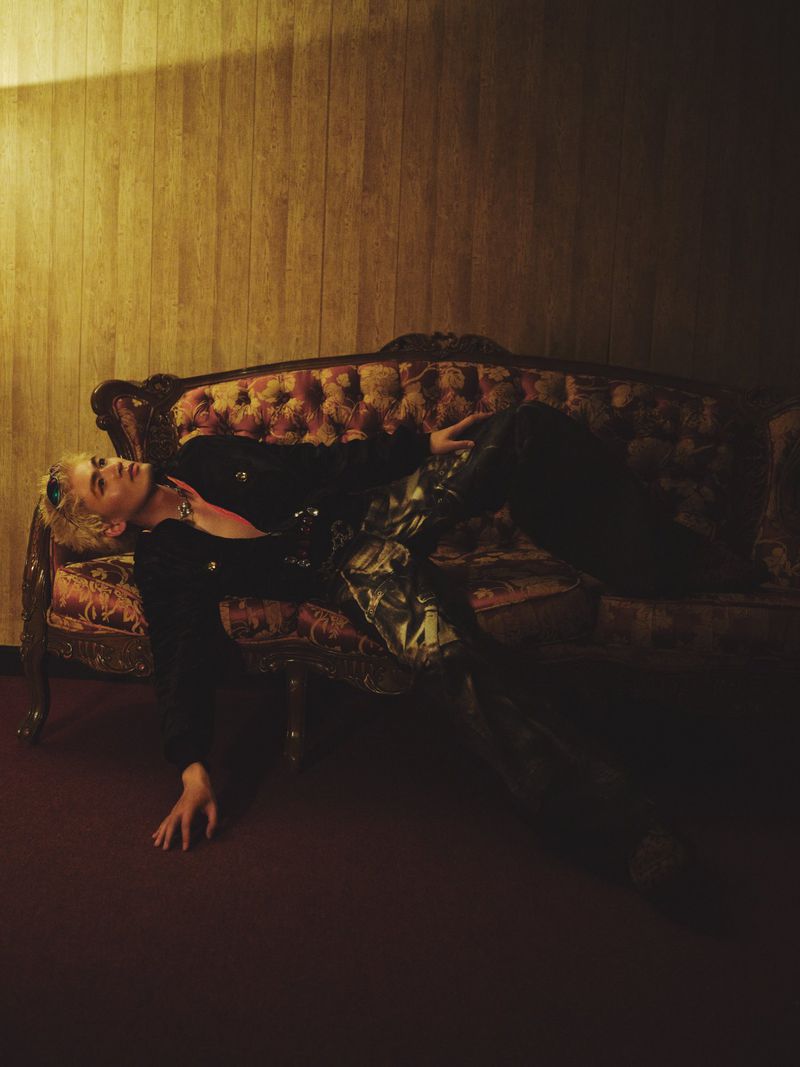 Model with blonde hair reclining dramatically on a vintage tufted sofa against a wood-paneled wall in warm, low lighting.