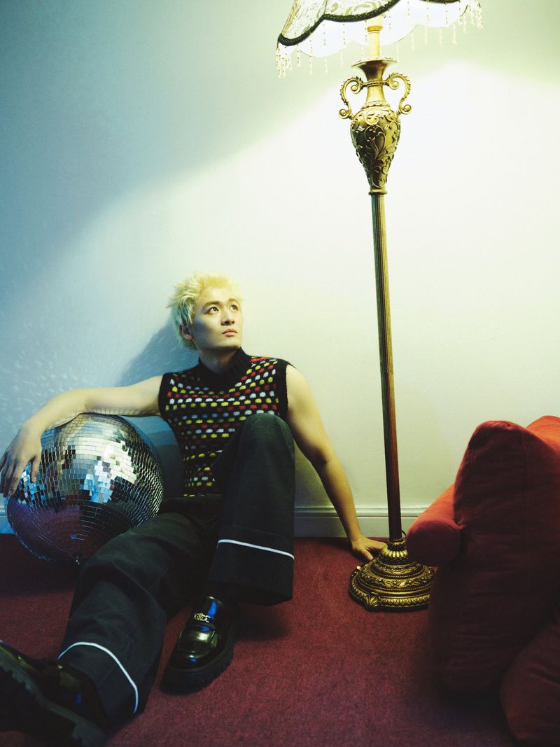 Man with bleached blonde hair in a patterned knit vest sitting on a red carpet beside a standing floor lamp and a disco ball.