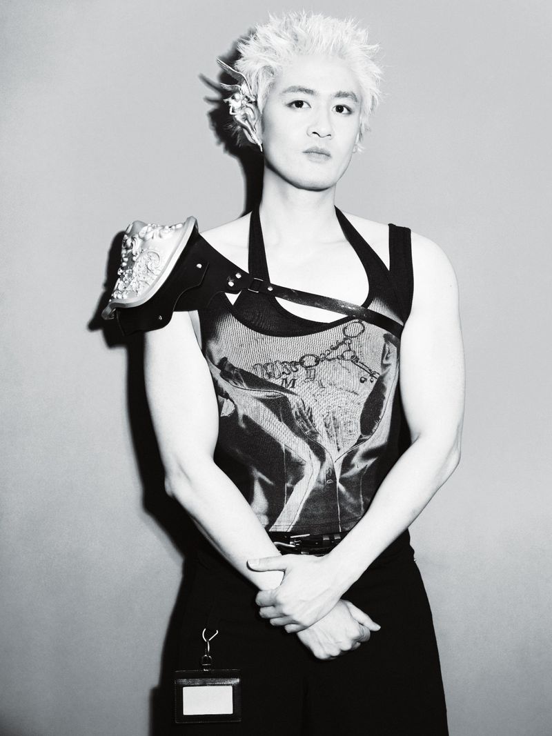 Black and white portrait of an East Asian man with spiky blonde hair wearing avant-garde armor-strap apparel against a studio wall.