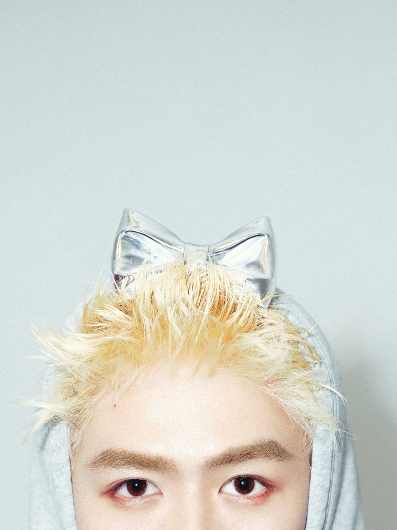 Close-up of a person with spiky bleached blonde hair wearing a silver metallic bow and grey hoodie against a pale studio backdrop.