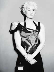 Black and white portrait of an East Asian man with spiky blonde hair wearing avant-garde armor-strap apparel against a studio wall.
