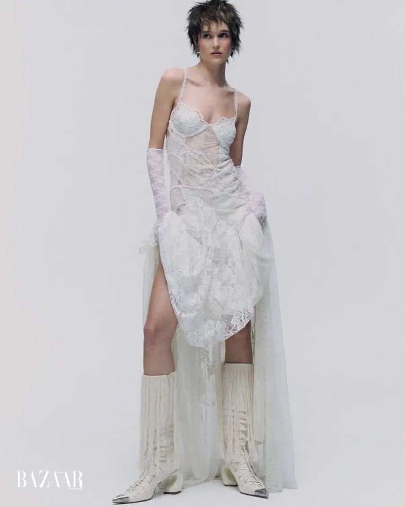 Model with short dark hair wearing a sheer white lace gown, lace gloves, and white fringe boots against a seamless studio background.