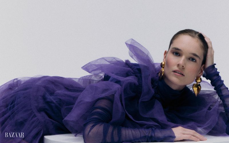 A model in a voluminous purple tulle gown and large gold statement earrings reclines against a light studio backdrop.