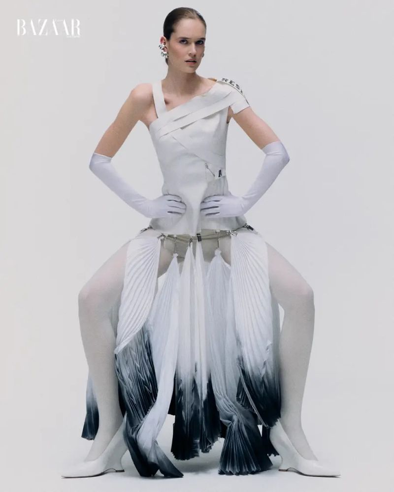 Fashion model in a structured white dress, opera gloves, and dip-dyed pleated skirt posing against a white studio background.